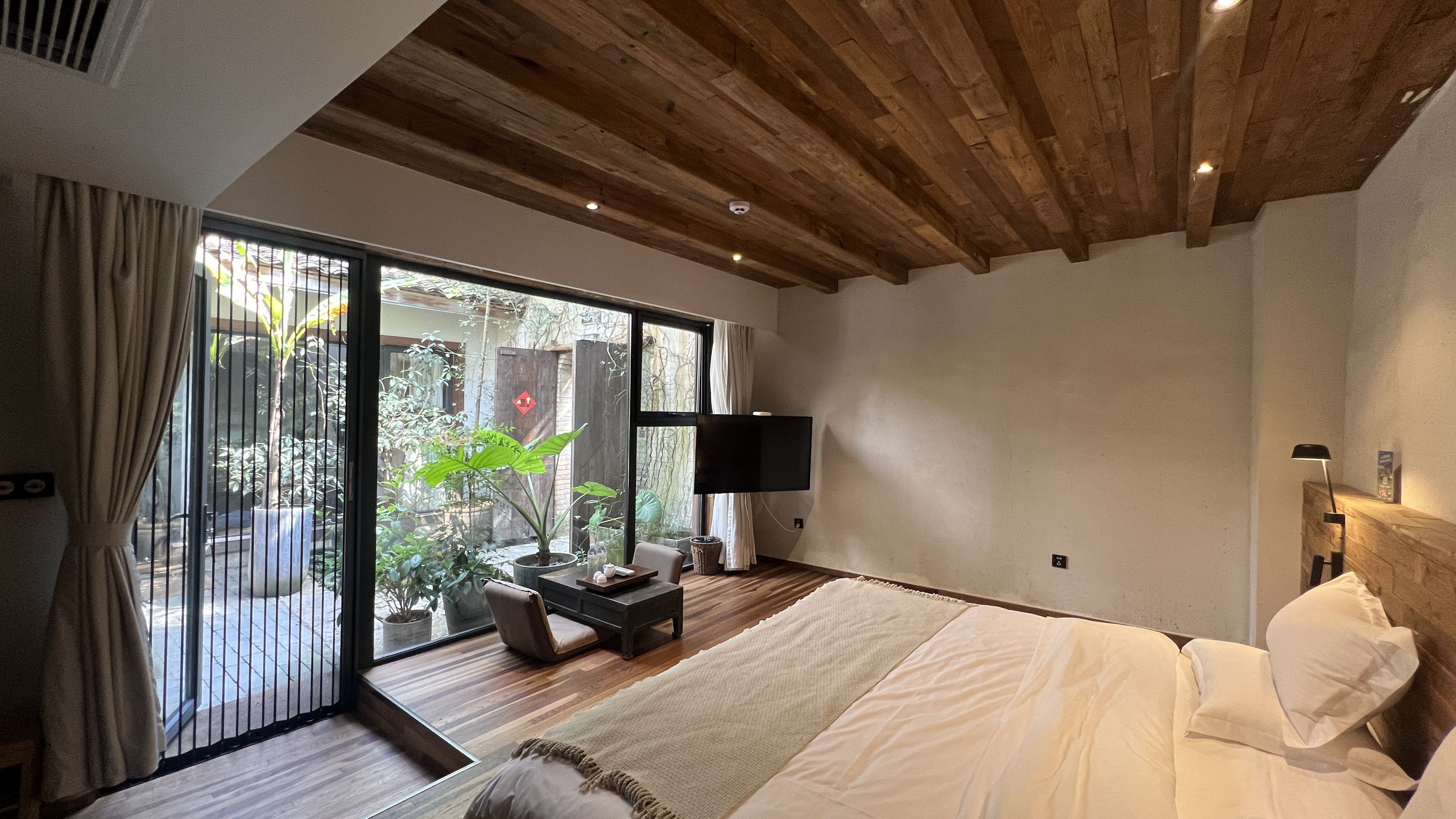 A bedroom with a large bed, a TV mounted on the wall, and a view of a garden through a sliding door.