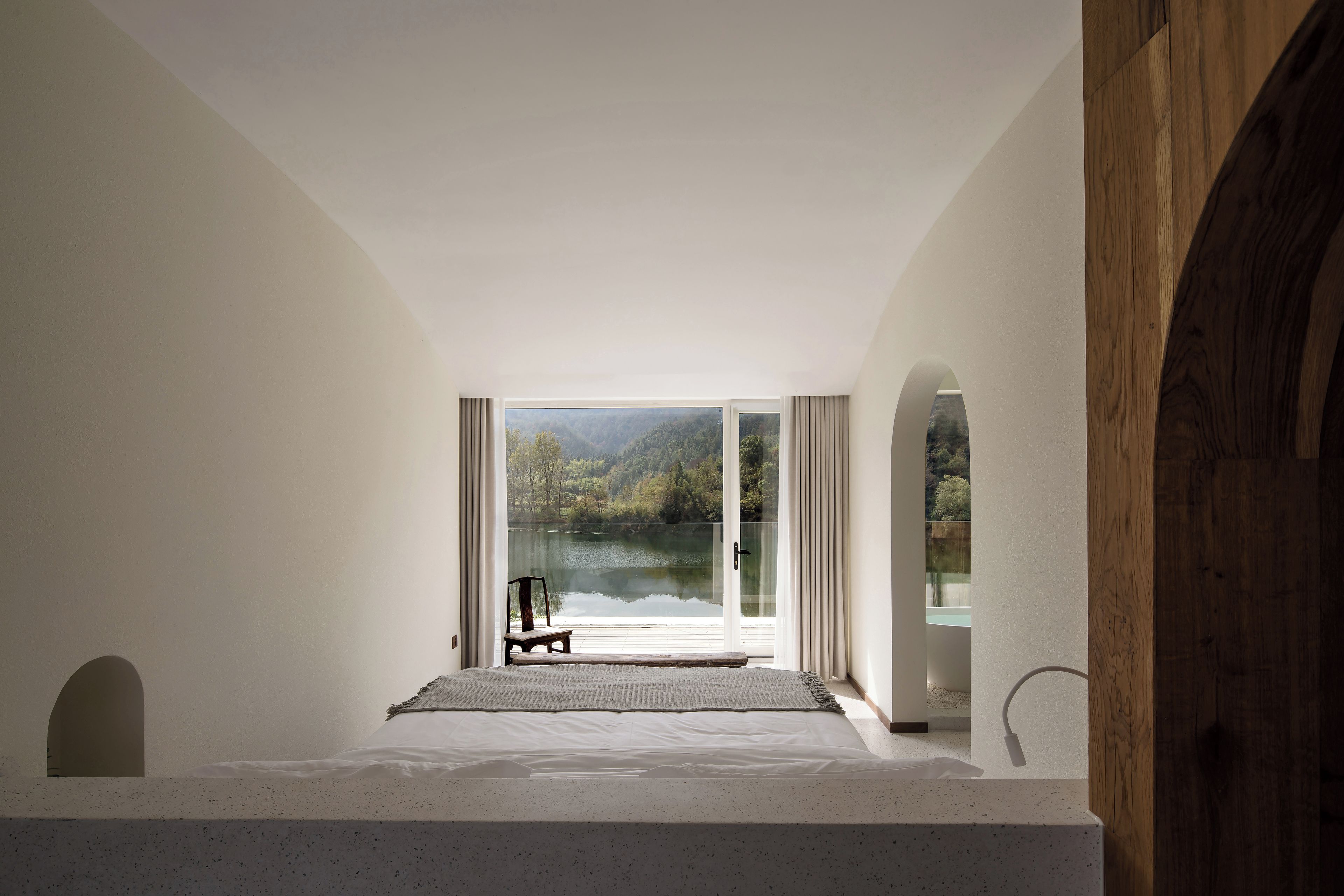 Un dormitorio con un ventanal grande, una cama, una silla y vistas a un lago y un bosque.