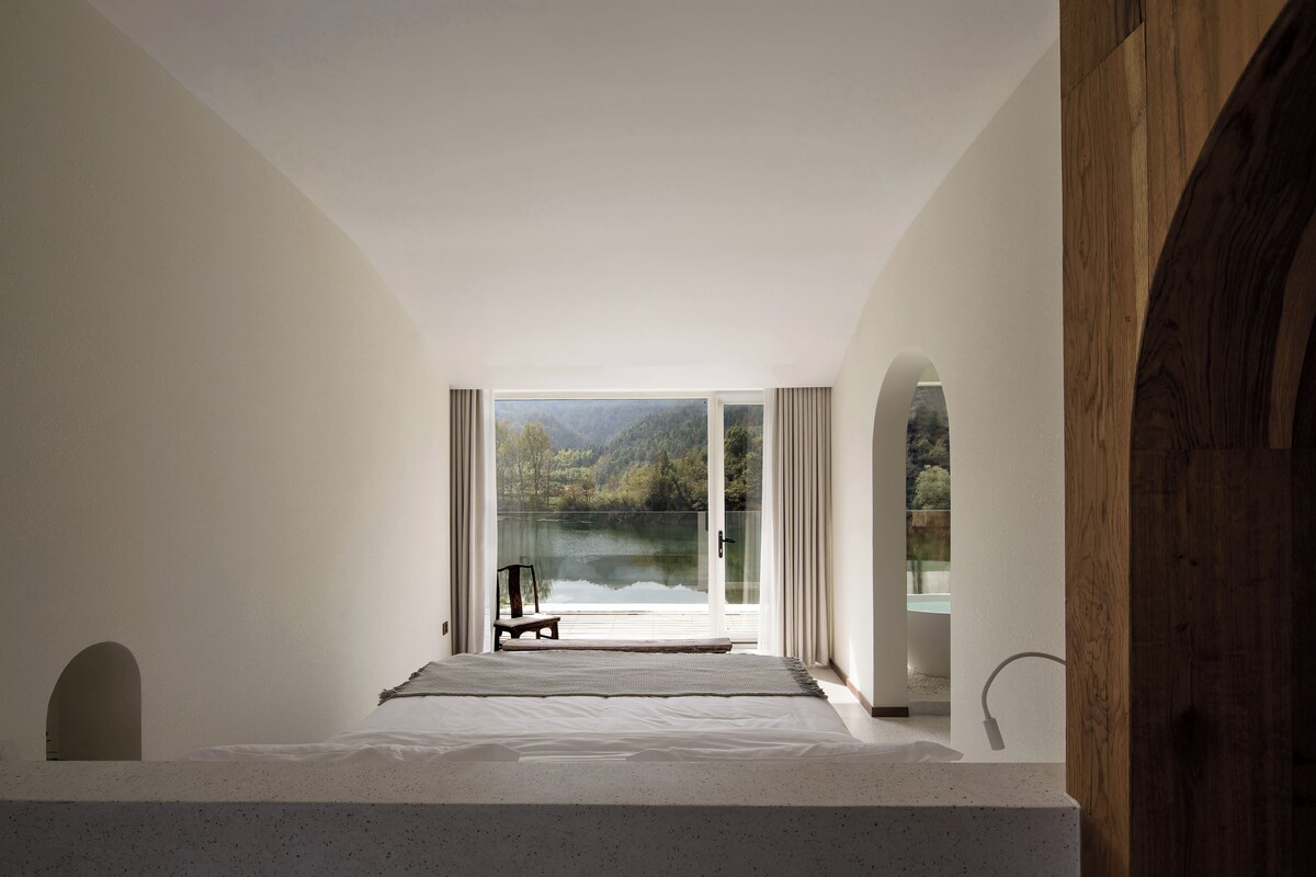 A bedroom with a large window, a bed, a chair, and a view of a lake and forest.