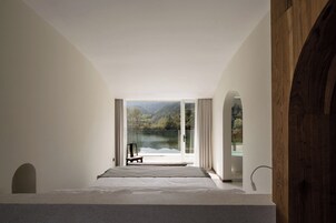 Quarto com uma janela grande, cama, poltrona e vista para um lago e uma floresta.