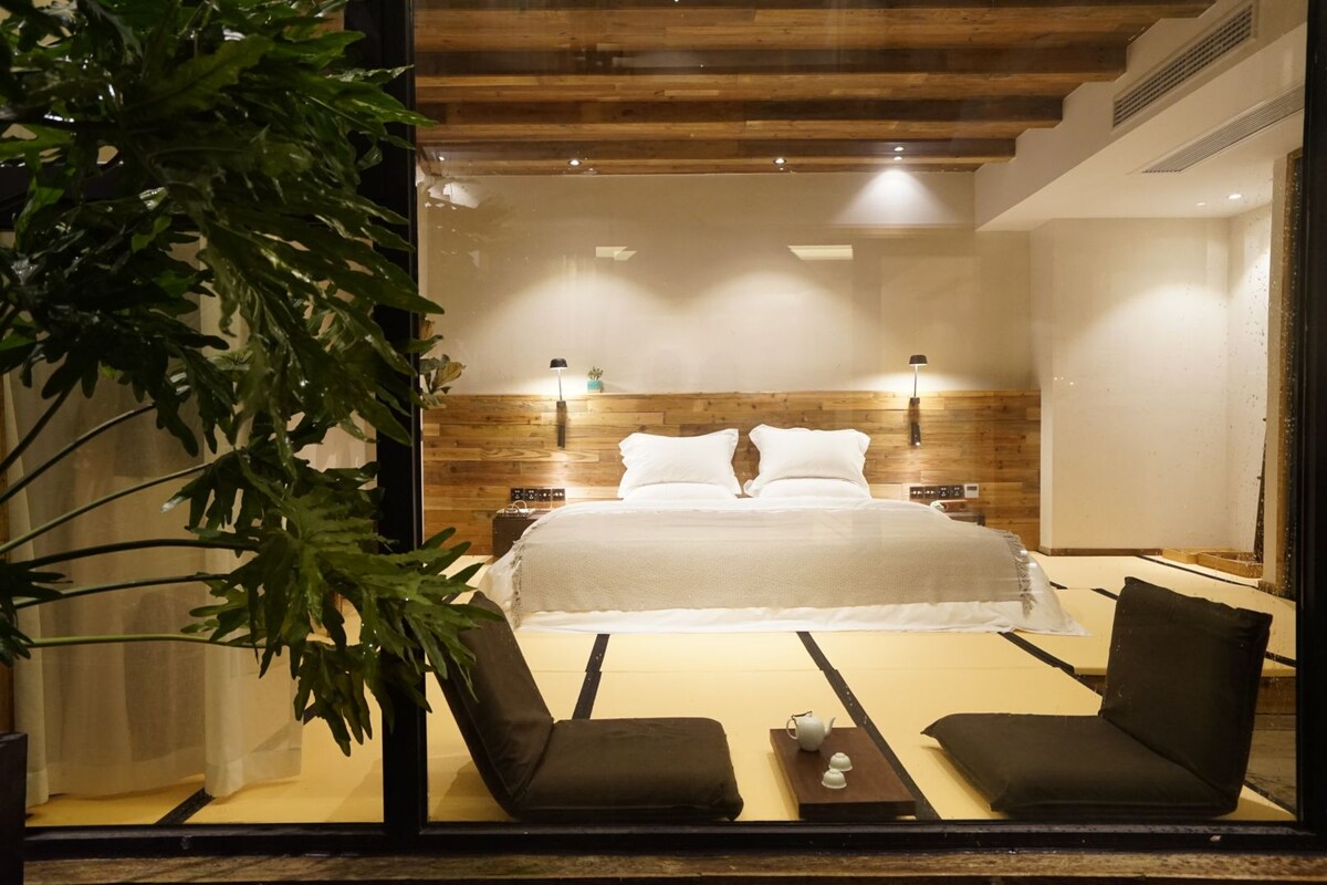 A modern bedroom with a large bed, two bedside tables, a nightstand, a plant, and a seating area with a table and chairs.