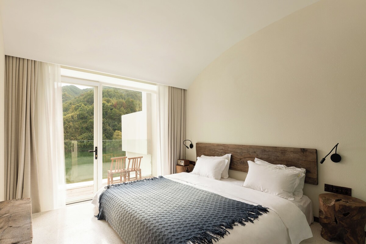 A hotel room with a large bed, a wooden headboard, a view of a forest through a sliding glass door, and a small table with a chair.