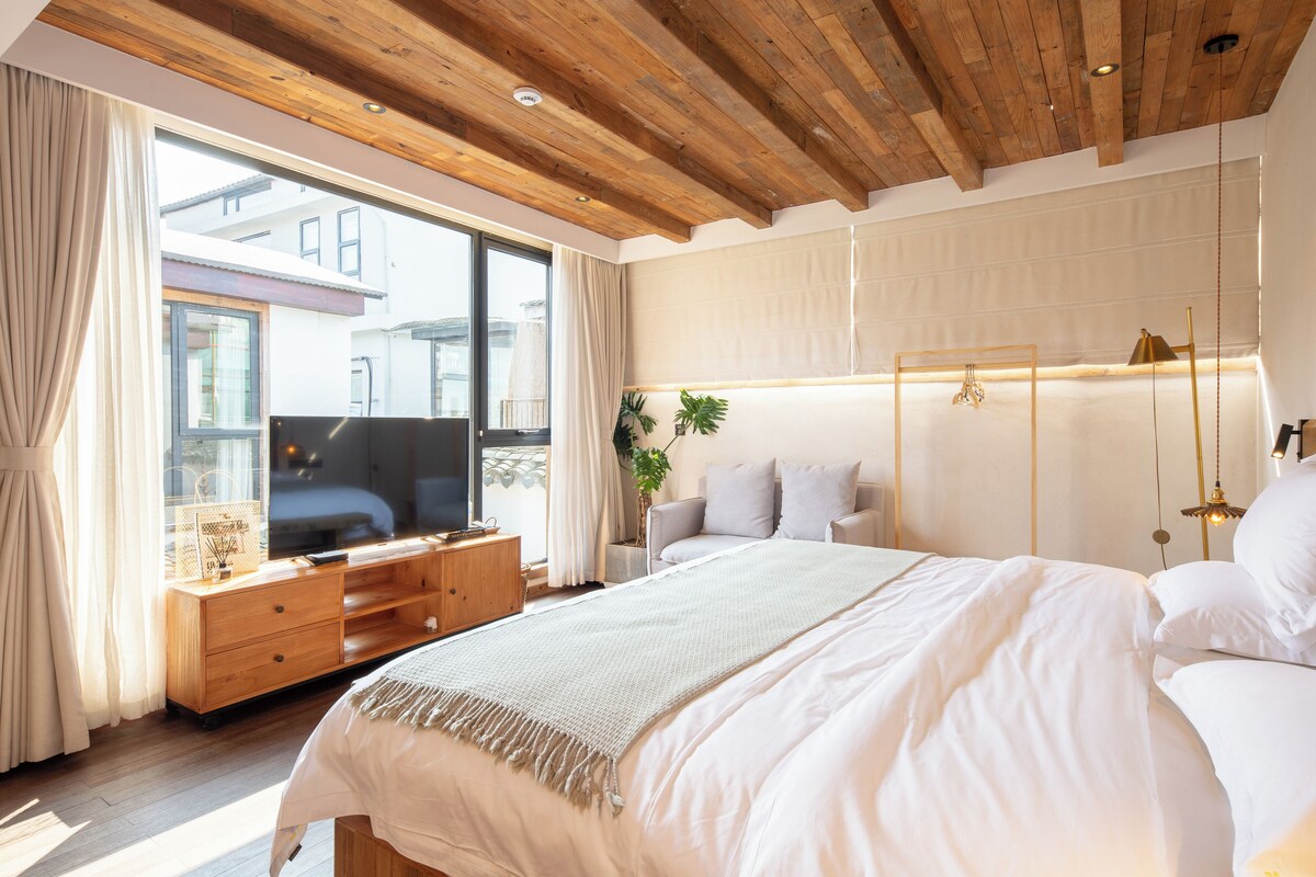 A modern bedroom with a wooden ceiling, a large bed, a TV on a wooden dresser, and a view of buildings outside.