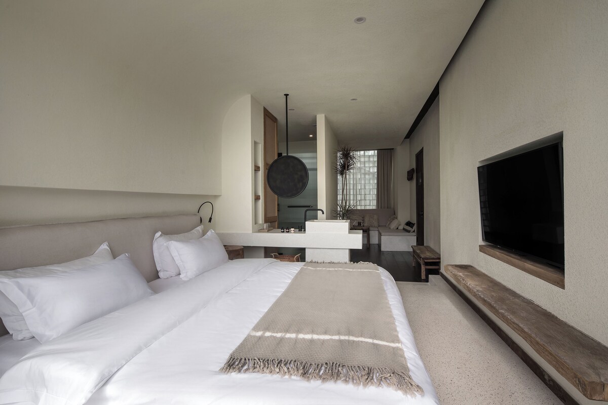 A modern bedroom with a large bed, a TV mounted on the wall, and a bathroom visible through an open door.