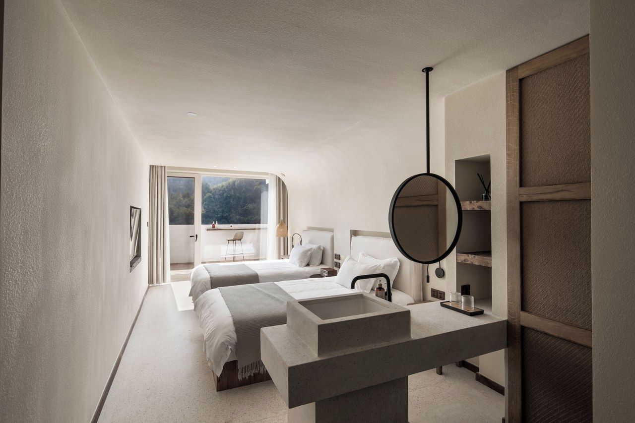 A modern hotel room with two beds, a large window, a round mirror, and a built-in desk.