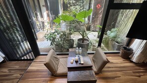 A room with a wooden floor, a sofa, a table with bottles and a remote control, and a view of an outdoor garden through a glass door. - ZHU85 (Lishui)