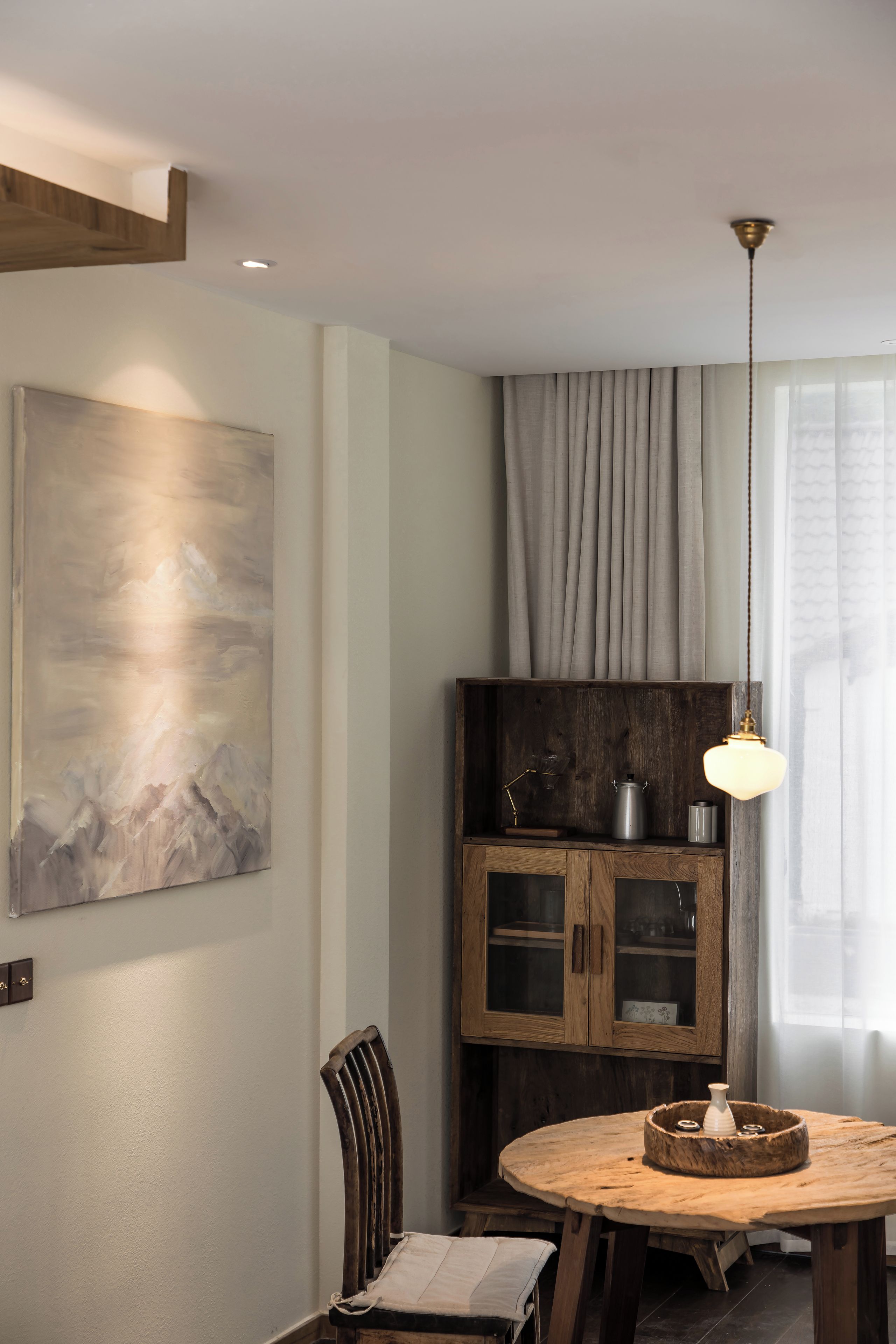 A dining area with a round wooden table, a chair, a pendant light, a cabinet with glass doors, and a painting on the wall.