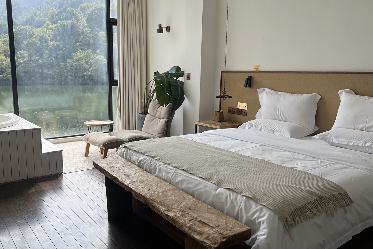 A modern bedroom with a large bed, a bench, a small table, and a view of a forest through a window.