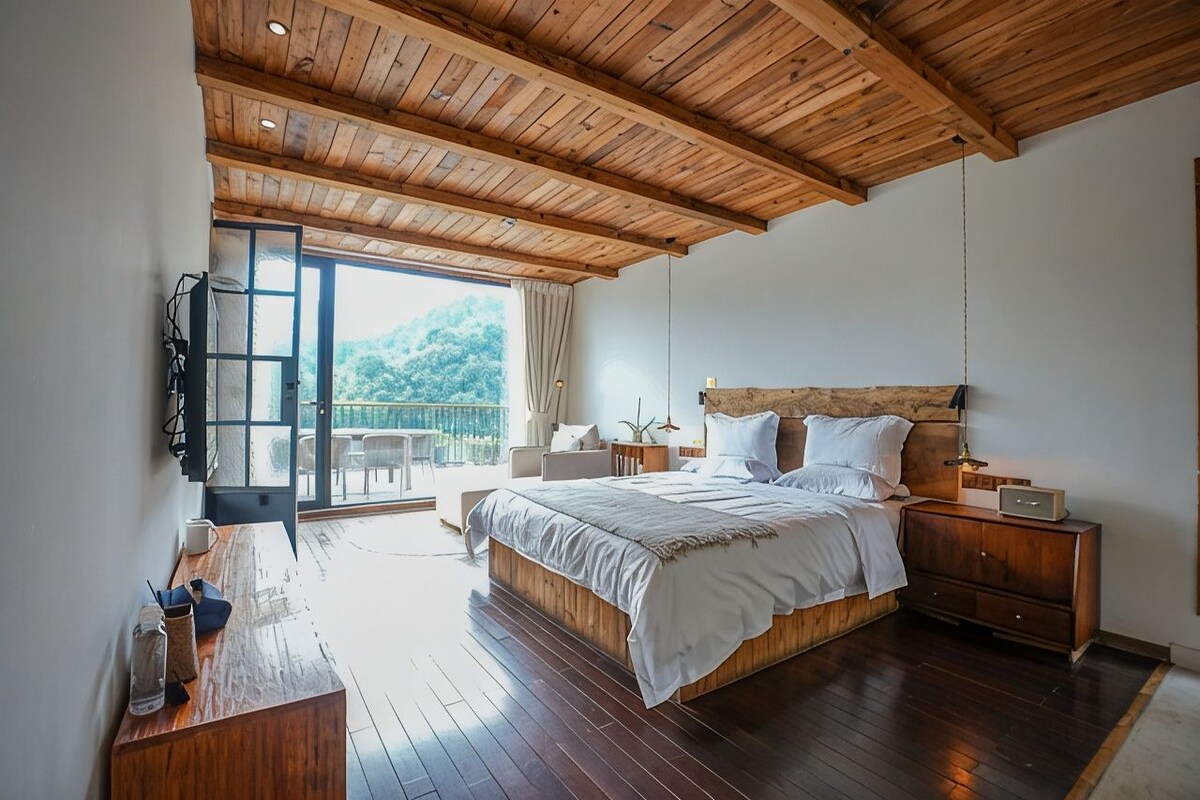 A bedroom with a wooden ceiling, a large bed, a nightstand, a bedside table, and a view of the outdoors.