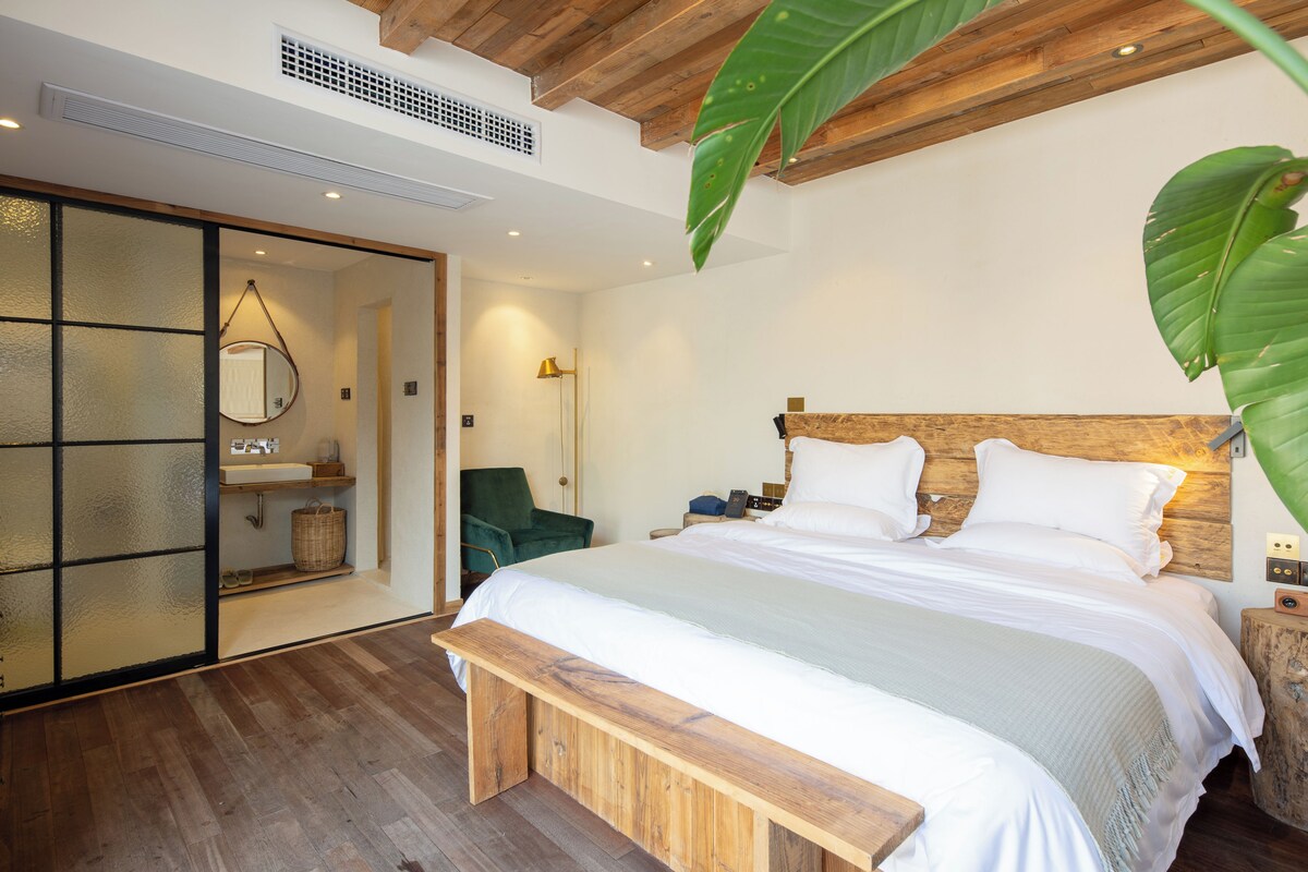 A bedroom with a wooden headboard, a large bed, a bench, a green chair, a wooden nightstand, and a mirror on the wall.