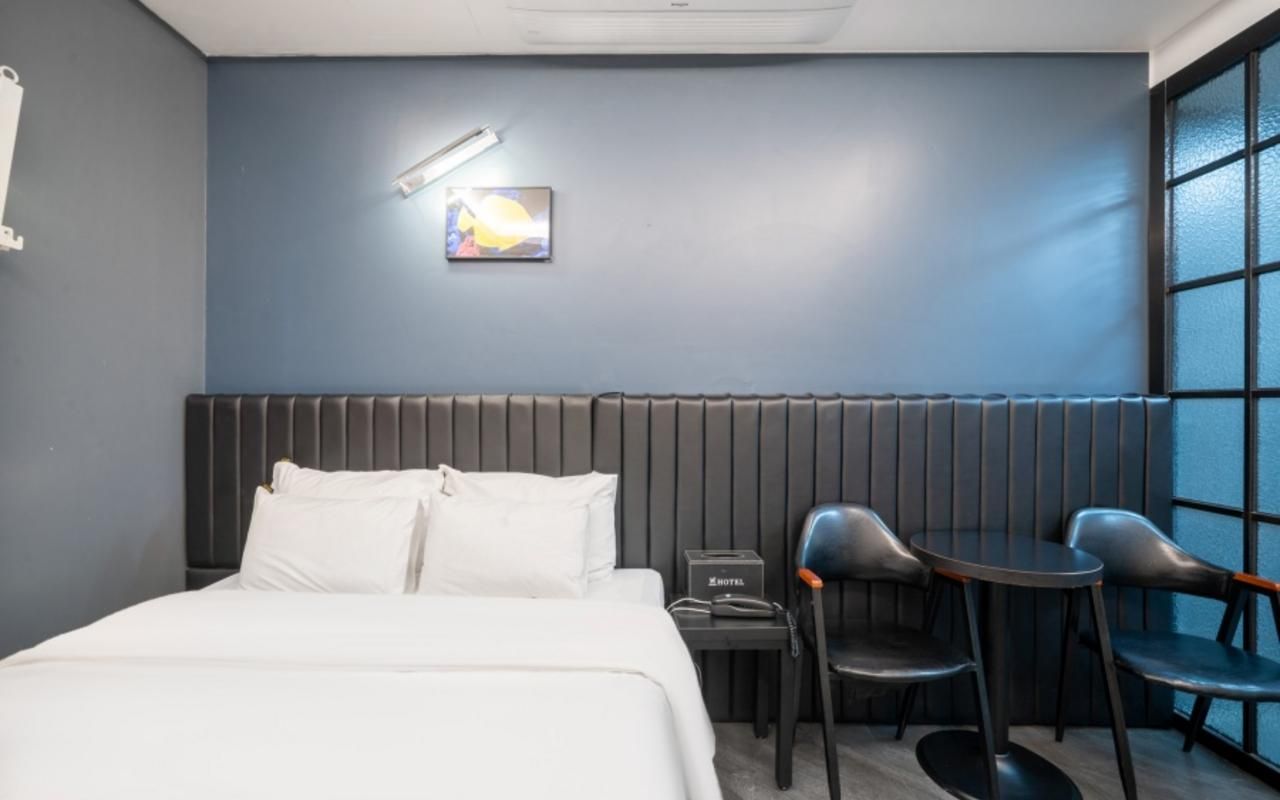 Habitación de hotel con cama, escritorio, dos sillas, mesita de noche, lámpara de pared y ventana con persianas.