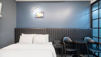 Habitación de hotel con cama, escritorio, dos sillas, mesita de noche, lámpara de pared y ventana con persianas.