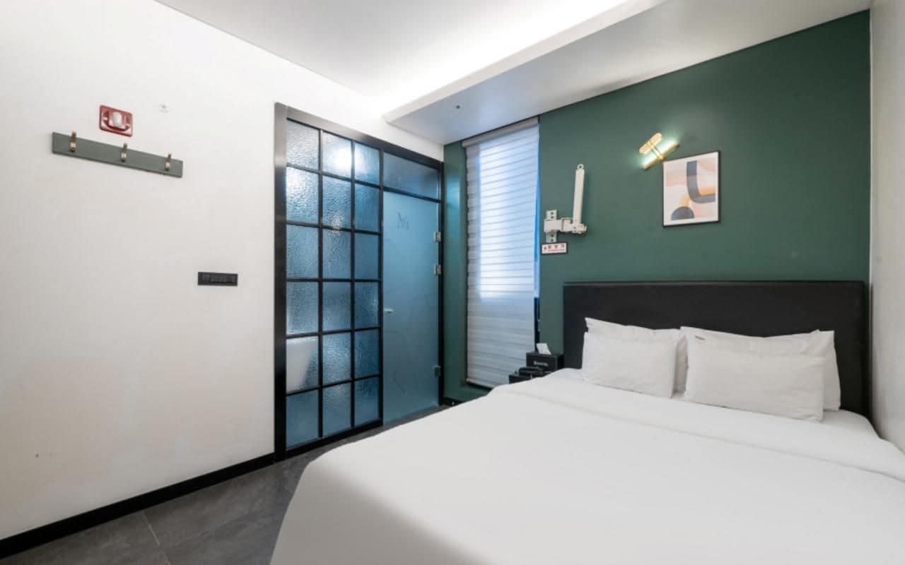 A hotel room with a bed, a door, a window with blinds, a wall-mounted light, and a small table with a phone.