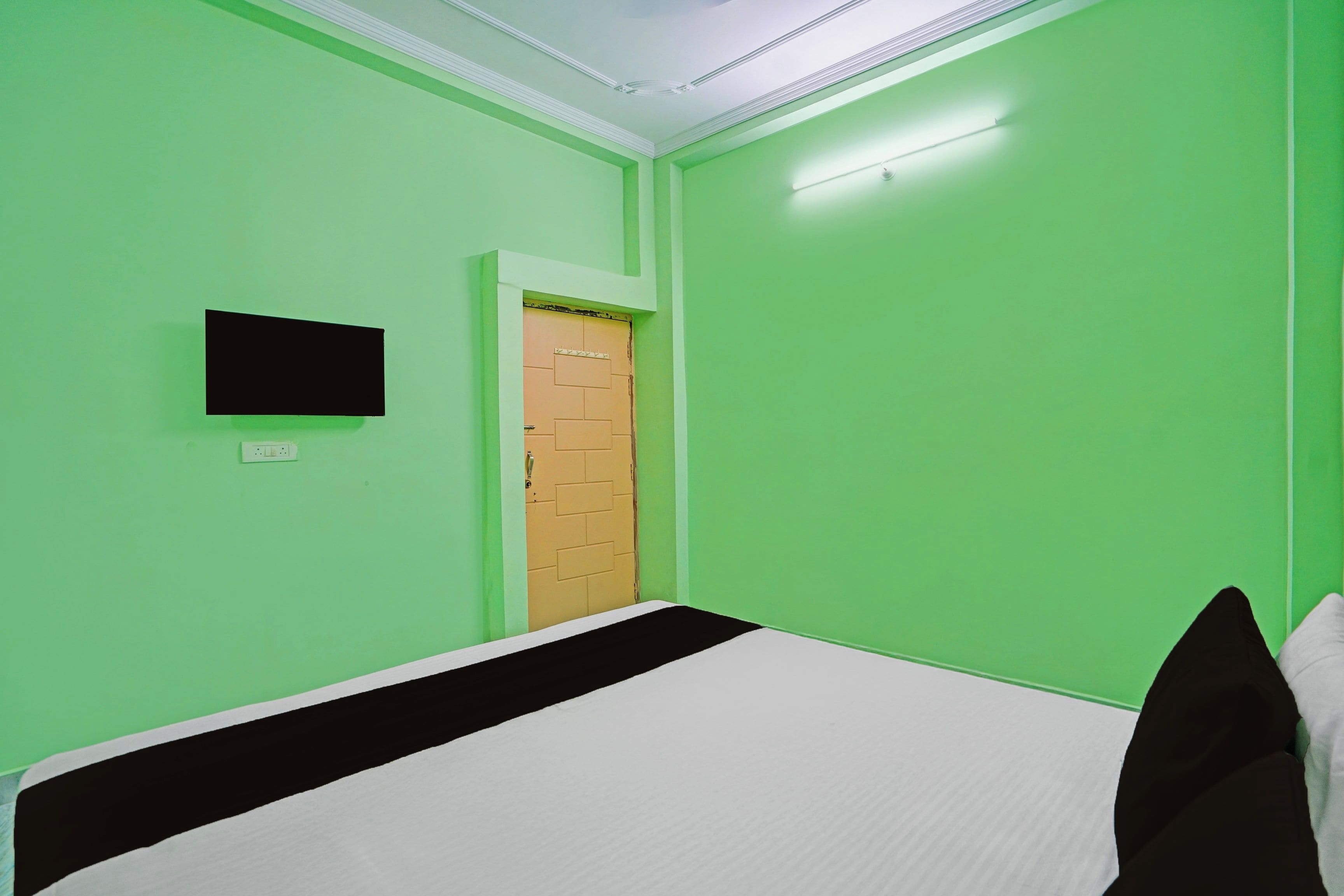Ein Zimmer mit grünen Wänden, einem Bett mit weißer und schwarzer Bettwäsche, einem an der Wand montierten Fernseher und einer Tür mit einem Holzpaneel.