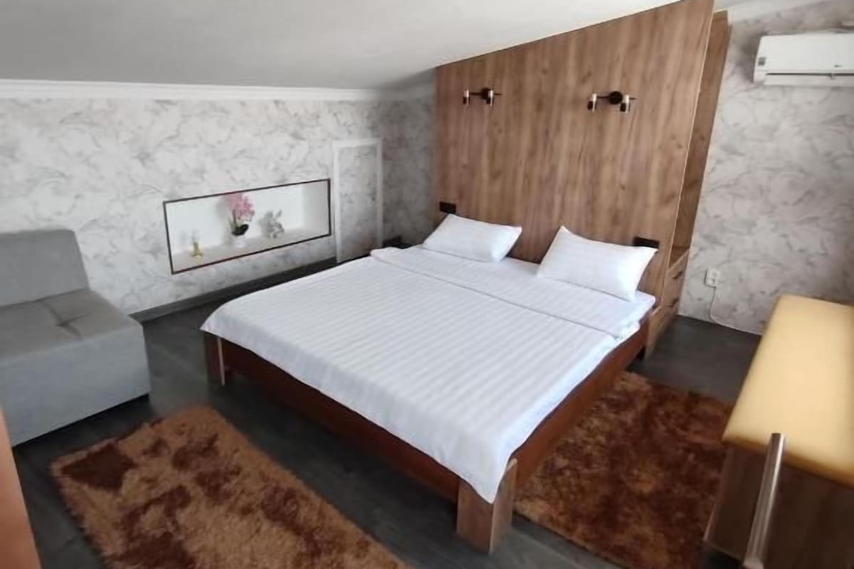 Un dormitorio con cabecera de madera, cama con ropa de cama blanca, sofá gris y alfombra marrón.