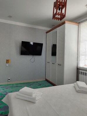 A hotel room with a bed, a television mounted on the wall, a wardrobe, and a window with blinds.