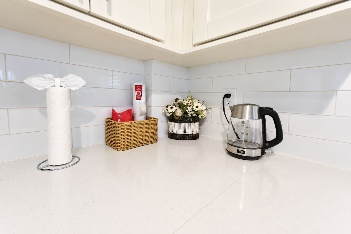 A kitchen with white tiles, a paper towel holder, a basket with a red item, a vase with flowers, and a kettle.