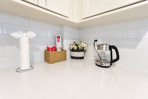 A kitchen with white tiles, a paper towel holder, a basket with a red item, a vase with flowers, and a kettle.
