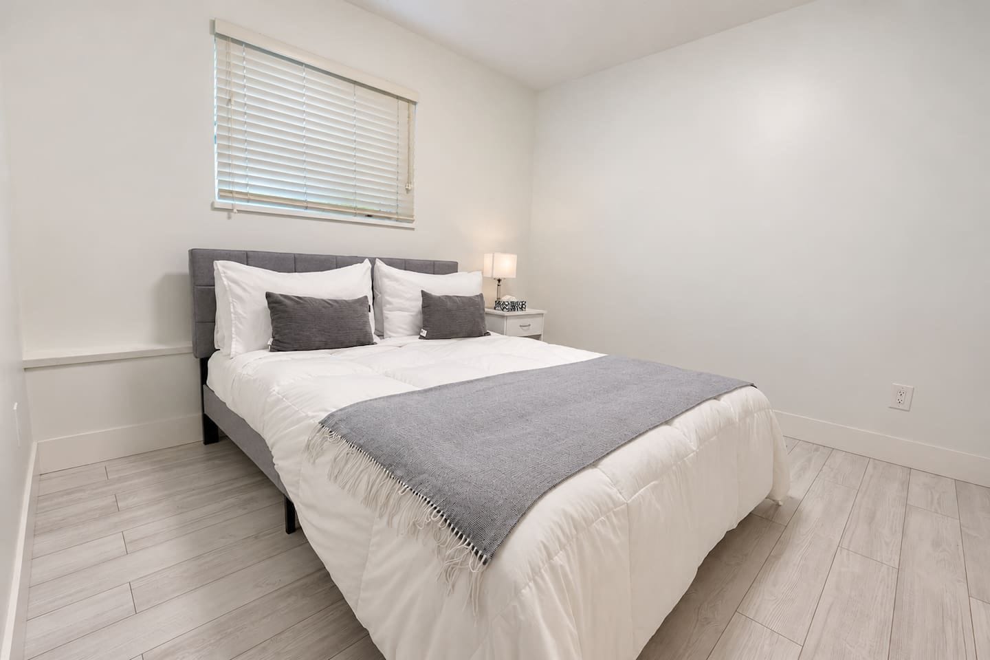 A bedroom with a bed, bedside table, lamp, and a window with blinds.