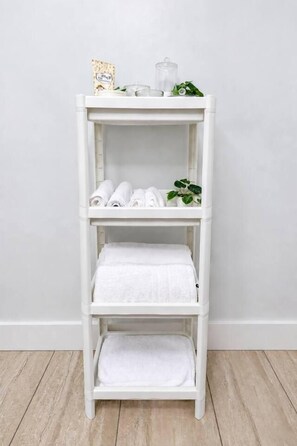 A white, three-tiered shelf unit with folded white towels on the top and bottom shelves, and a clear glass container on the middle shelf.