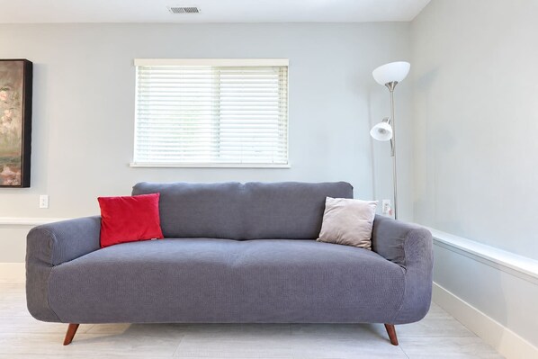 A grey sofa with two cushions, a floor lamp, and a window with blinds.