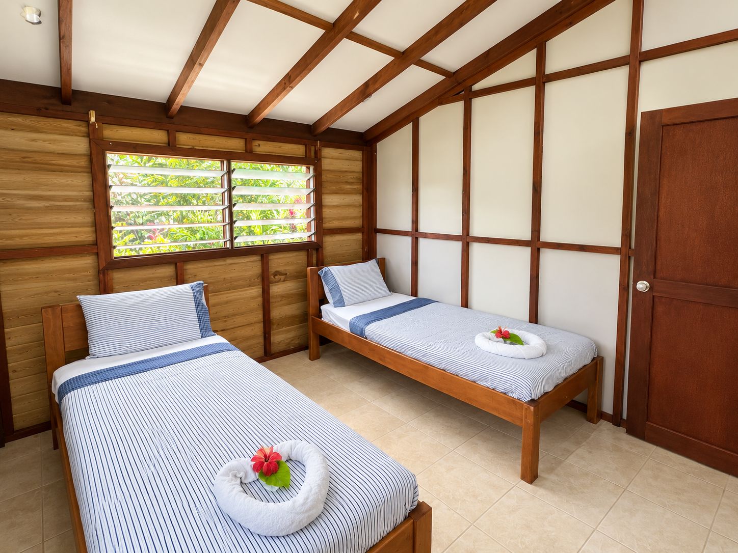 A room with two beds, wooden beams on the ceiling, a window with a view of greenery, and a door.