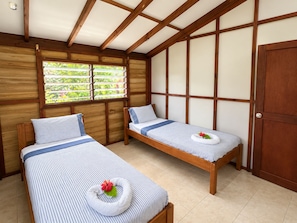 A room with two beds, wooden beams on the ceiling, a window with a view of greenery, and a door.
