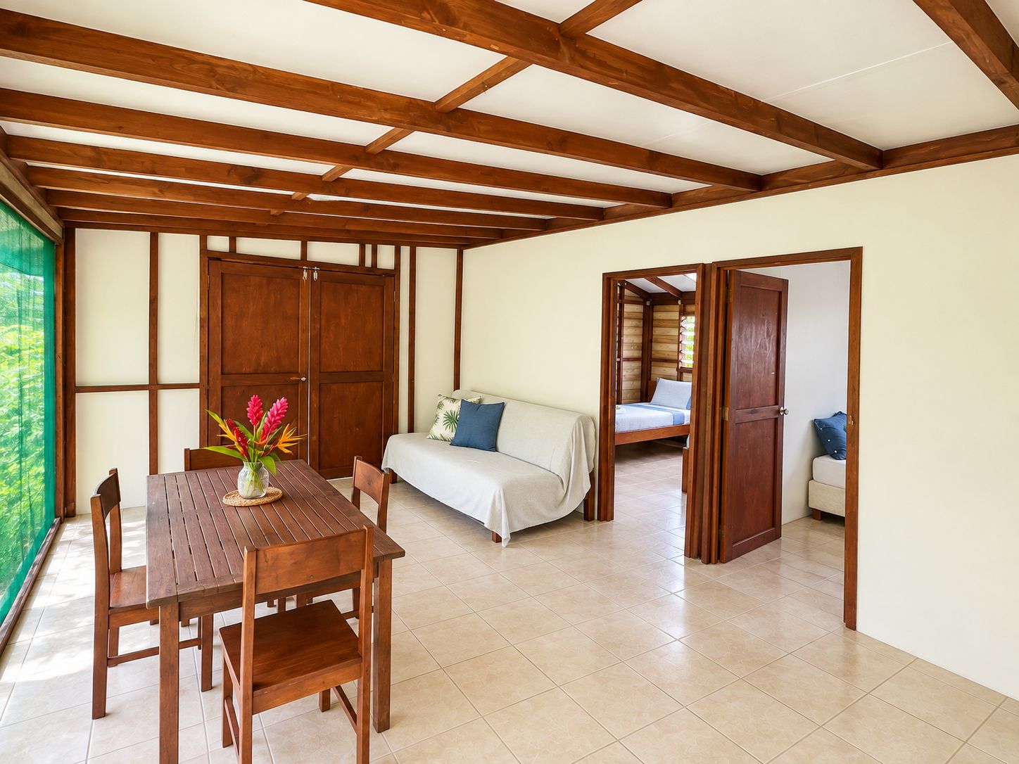 A room with a wooden table and chairs, a sofa, and a bed with a view of greenery outside.