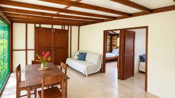 A room with a wooden table and chairs, a sofa, and a bed with a view of greenery outside.