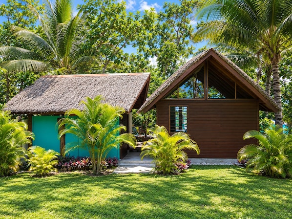 A small wooden house with a thatched roof, surrounded by lush tropical vegetation.