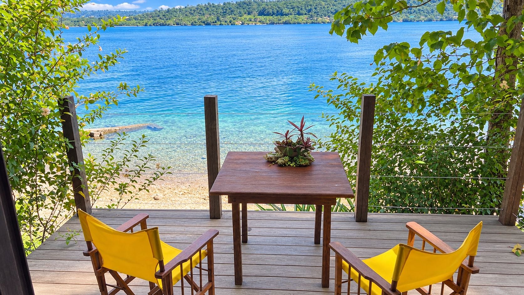 A wooden deck with a table and two yellow chairs, overlooking a clear blue lake.