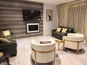 Een moderne woonkamer met een flatscreen televisie aan een grijze bakstenen muur, een groene leren bank met gekleurde kussens en twee ronde fauteuils met gouden details.