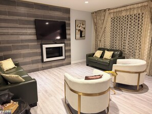 A modern living room with a flat-screen TV mounted on a brick-patterned wall, a leather sofa, two armchairs, and a glass coffee table. - Nolan Hill Mountain Gateway | Spacious 4BR Home (Calgary)