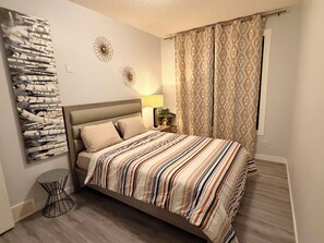 Une chambre à coucher avec un lit, une œuvre d’art accrochée au mur, une table de chevet avec une lampe et des rideaux à motifs.