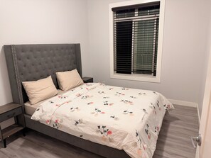 A bedroom with a tufted headboard, a floral bedspread, a nightstand, and a window with blinds. - Nolan Hill Mountain Gateway | Spacious 4BR Home (Calgary)