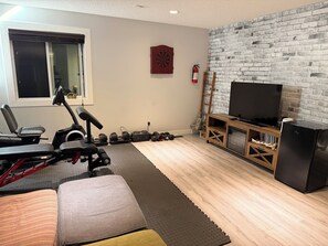 A room with a treadmill, exercise equipment, a television, and a brick wall. - Nolan Hill Mountain Gateway | Spacious 4BR Home (Calgary)