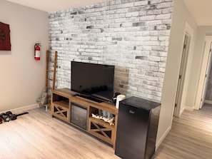 A living room with a brick wall, a flat-screen TV, wooden entertainment unit, a refrigerator, and a fire extinguisher. - Nolan Hill Mountain Gateway | Spacious 4BR Home (Calgary)