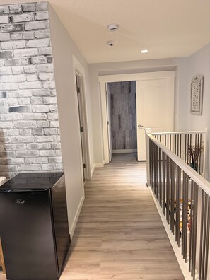A hallway with a brick wall, a black refrigerator, and a wooden floor leading to a door. - Nolan Hill Mountain Gateway | Spacious 4BR Home (Calgary)