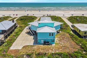 A coastal building with a steep roof, a staircase leading to the entrance, and a view of the beach and ocean in the background. - A Sight for Shore Eyes - Beachfront Home, Sleeps 10 (Please note: The third floor and garage are not available to guests.) (Port Aransas)