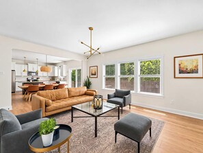 A spacious living room with a modern sofa, a coffee table, and a dining area with a kitchen in the background. - Lakeshore Retreat | Hot Tub • 5BR • Steps to Beach (South Haven)