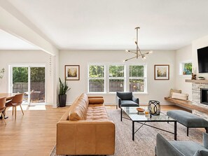 Ein geräumiges Wohnzimmer mit einer großen Couch, einem Couchtisch und einem Kamin. Mehrere Fenster sorgen für natürliches Licht und Ausblicke ins Freie.