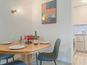 A dining area with a round table, chairs, and a bottle of wine. There is a painting on the wall and a kitchen in the background. - Domino B 38 by Interhome (Ovronnaz)