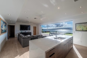 A modern kitchen with a large island, a dining area with a view of the sea, and a living room with a sofa and artwork. - The Panorama Beach House (Wailuku)