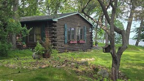 A rustic log cabin with a porch, surrounded by lush greenery and a body of water in the background. - Pine Beach Peninsula iconic property on Gull Lake. Truly one of a kind & special (City of East Gull Lake)