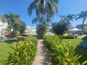 Caminho cercado por plantas tropicais que leva a uma área de piscina com espreguiçadeiras e guarda-sóis.