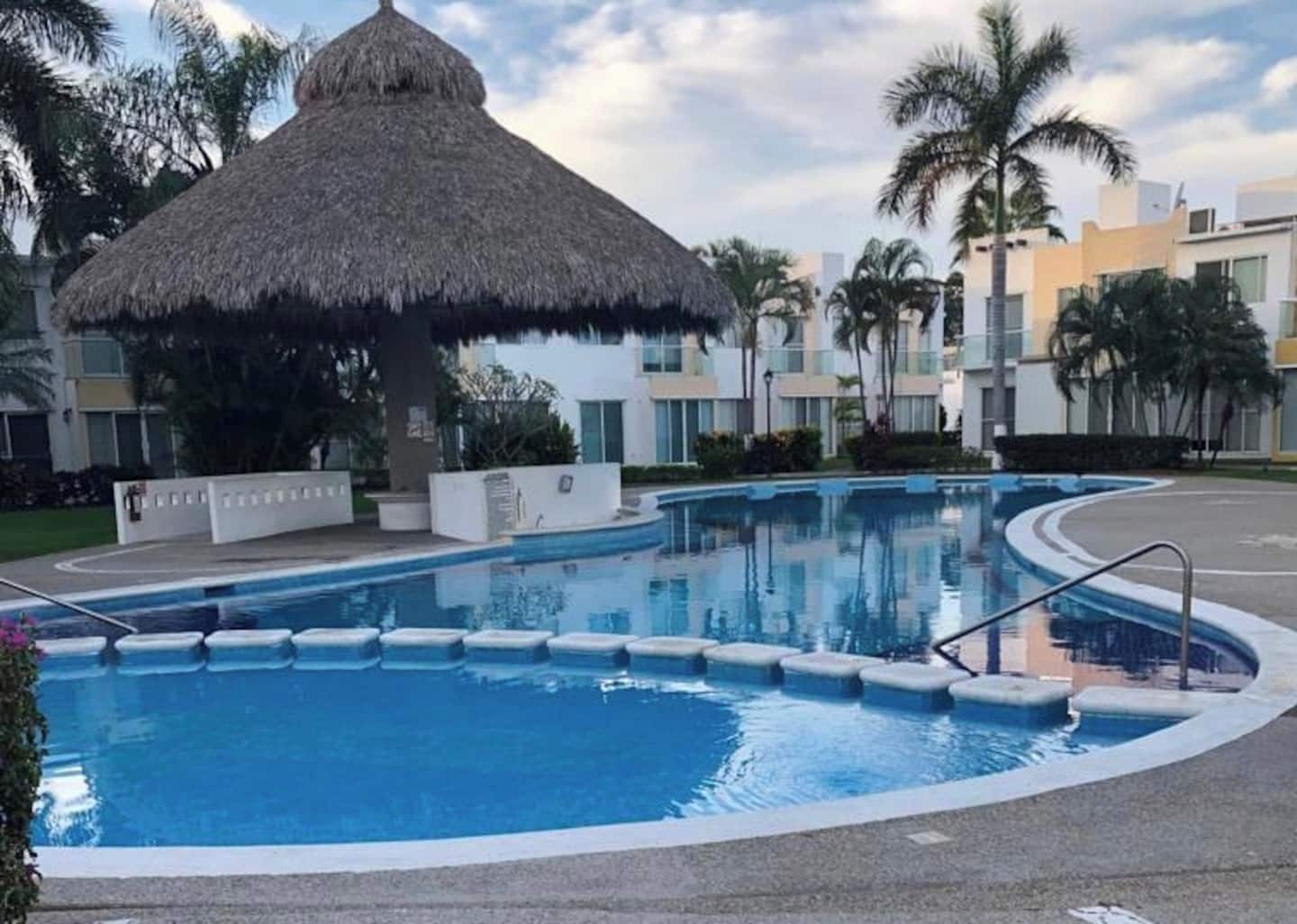 Una piscina con una estructura de techo de paja, rodeada de palmeras y edificios modernos.