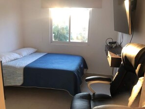 Quarto com cama, escrivaninha, cadeira e televisão.