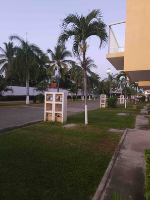 A well-maintained grassy area with palm trees, a paved walkway, and a modern building with a yellow facade. - Casa Diamante: private pool, paddle court, 24/7 security (Acapulco de Juárez)