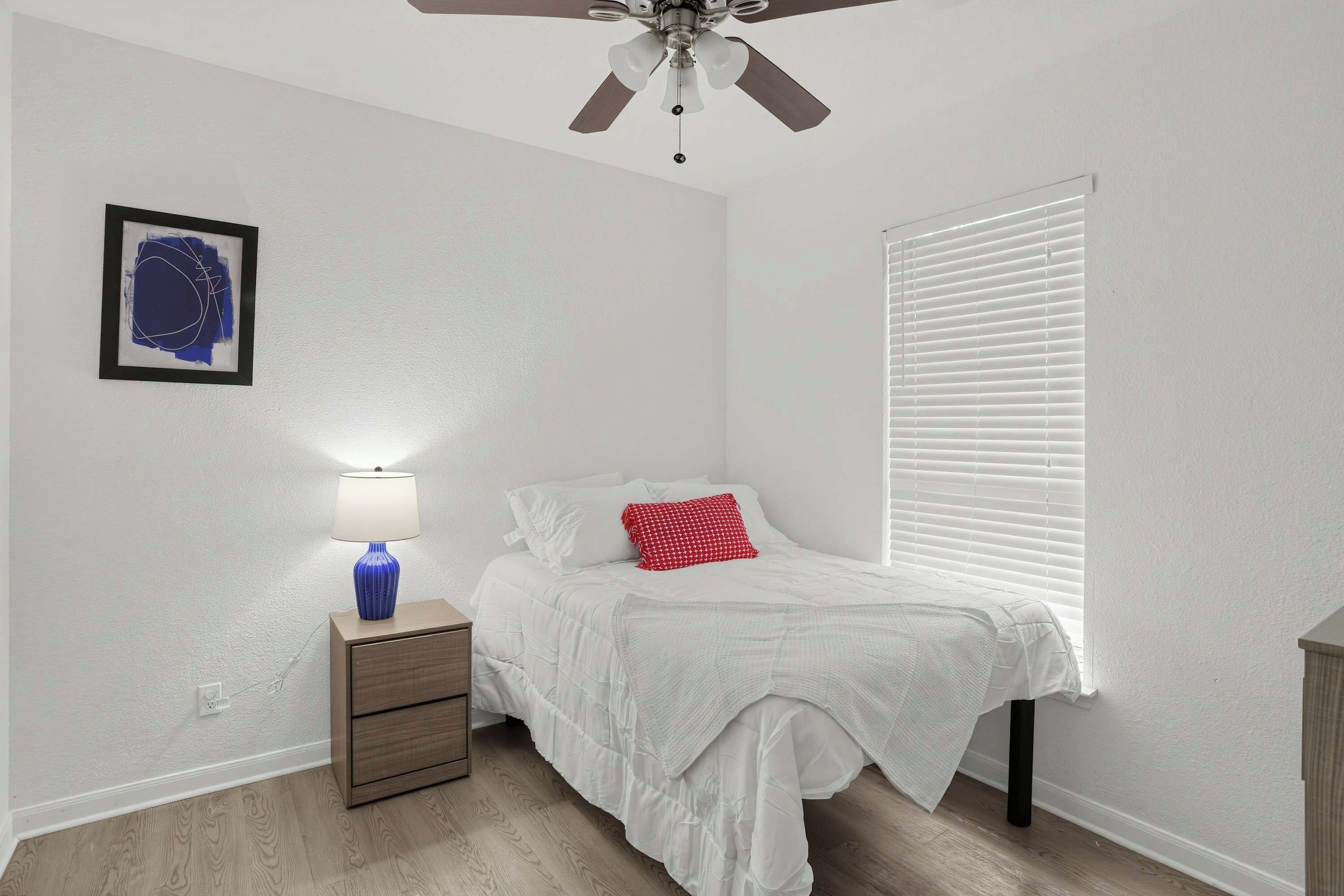 Une chambre à coucher comprenant un lit, une table de chevet, une lampe, un ventilateur de plafond et une fenêtre avec des stores.