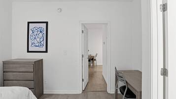 Un dormitorio con cama, cómoda, escritorio y silla. Hay una puerta que da a un comedor con mesa y sillas.
